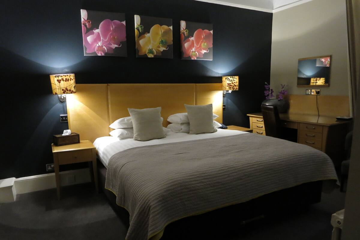 A hotel room with a large bed, two bedside tables, a nightstand, a desk, and a chair. There are three framed pictures on the wall above the bed.