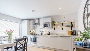 Una cocina moderna con gabinetes blancos, horno empotrado, microondas y estufa a gas. Hay un comedor con mesa y sillas, y un ventanal con persianas.