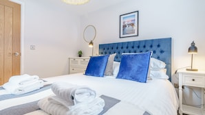Una cama bien hecha con ropa de cama azul y blanca, una mesita de noche blanca con una lámpara, y un cuadro en la pared.