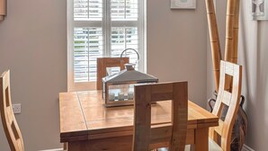 A dining area with a wooden table and chairs, a window with blinds, and a wall-mounted light fixture. - 4 bedroom accommodation in Sidmouth (Sidmouth)