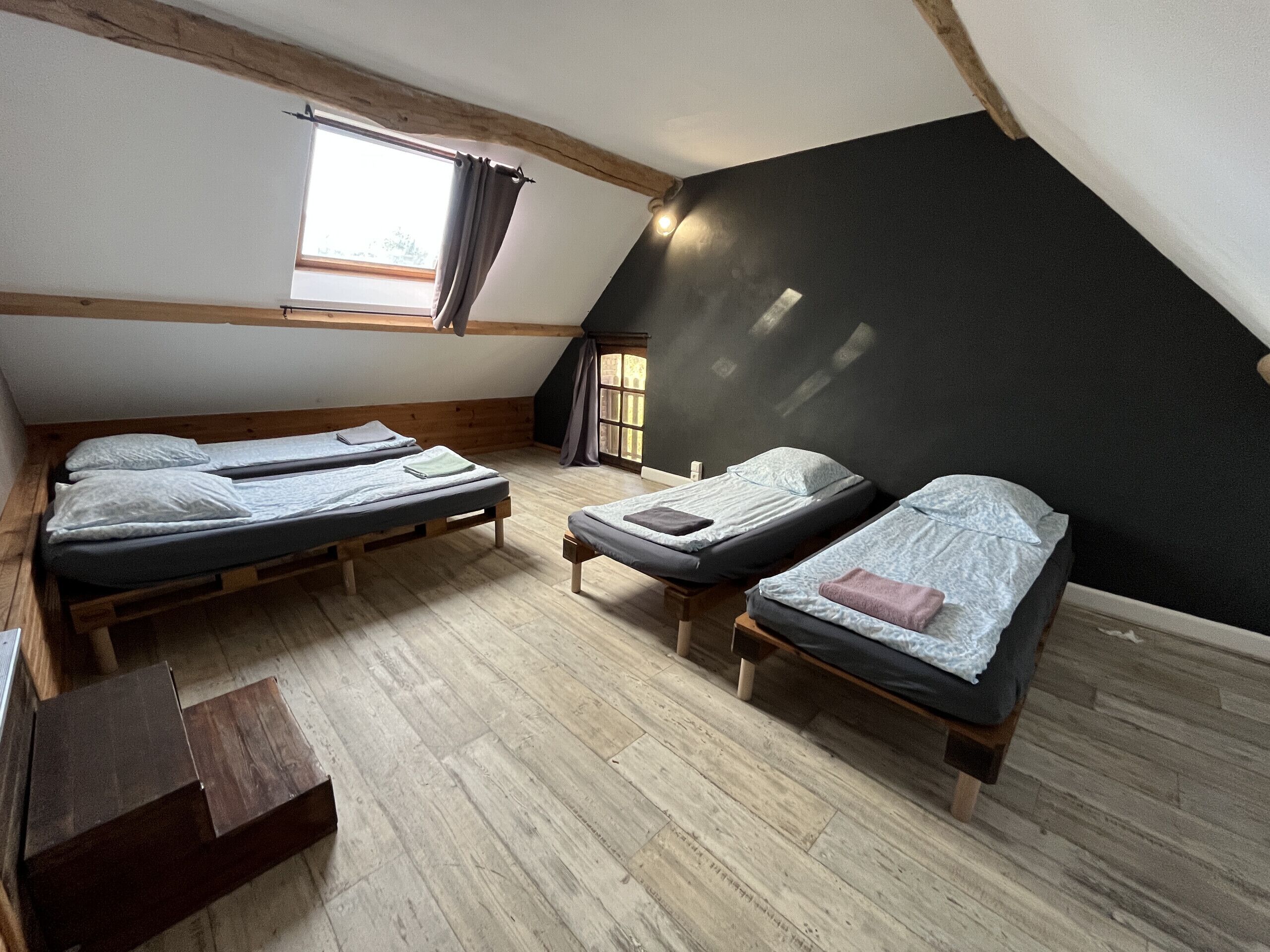 A dormitory room with three bunk beds, wooden flooring, a window with curtains, and a sloped ceiling.