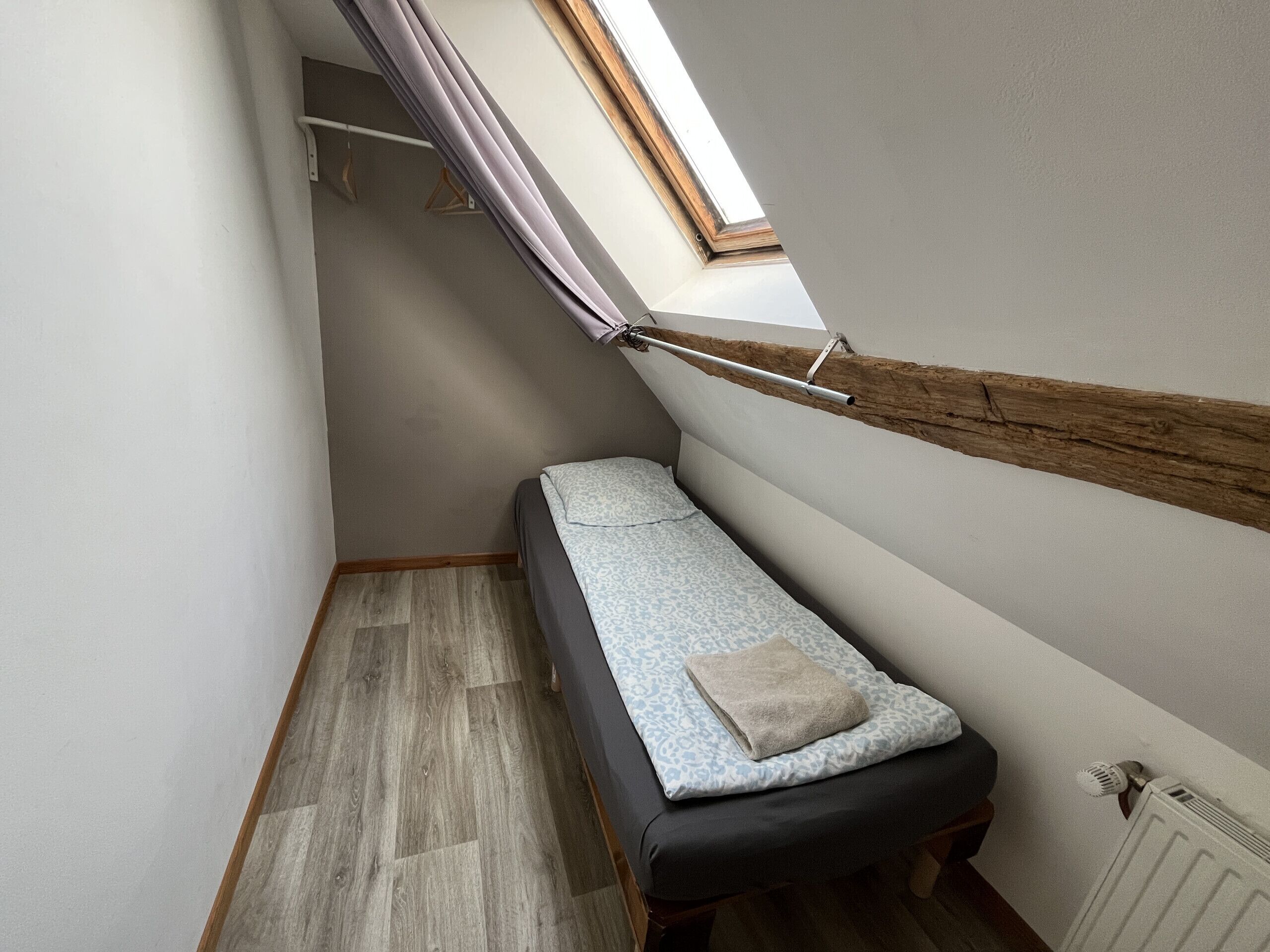 Een kleine, smalle kamer met een enkel bed, een houten balkenplafond en een dakraam.