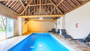 Piscina interior con techo de madera, suelo de baldosas y sillones de descanso a un lado.