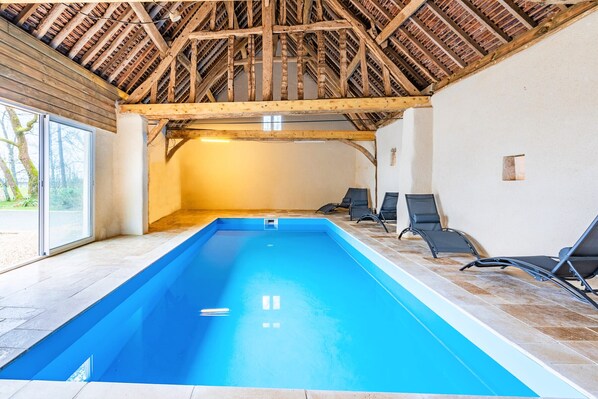 Piscina interior con techo de madera, suelo de baldosas y sillones de descanso a un lado.