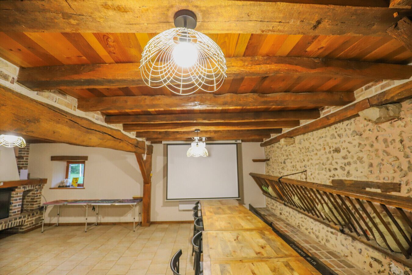 Een rustieke kamer met zichtbare houten balken, een open haard, een projectiescherm en een trap.