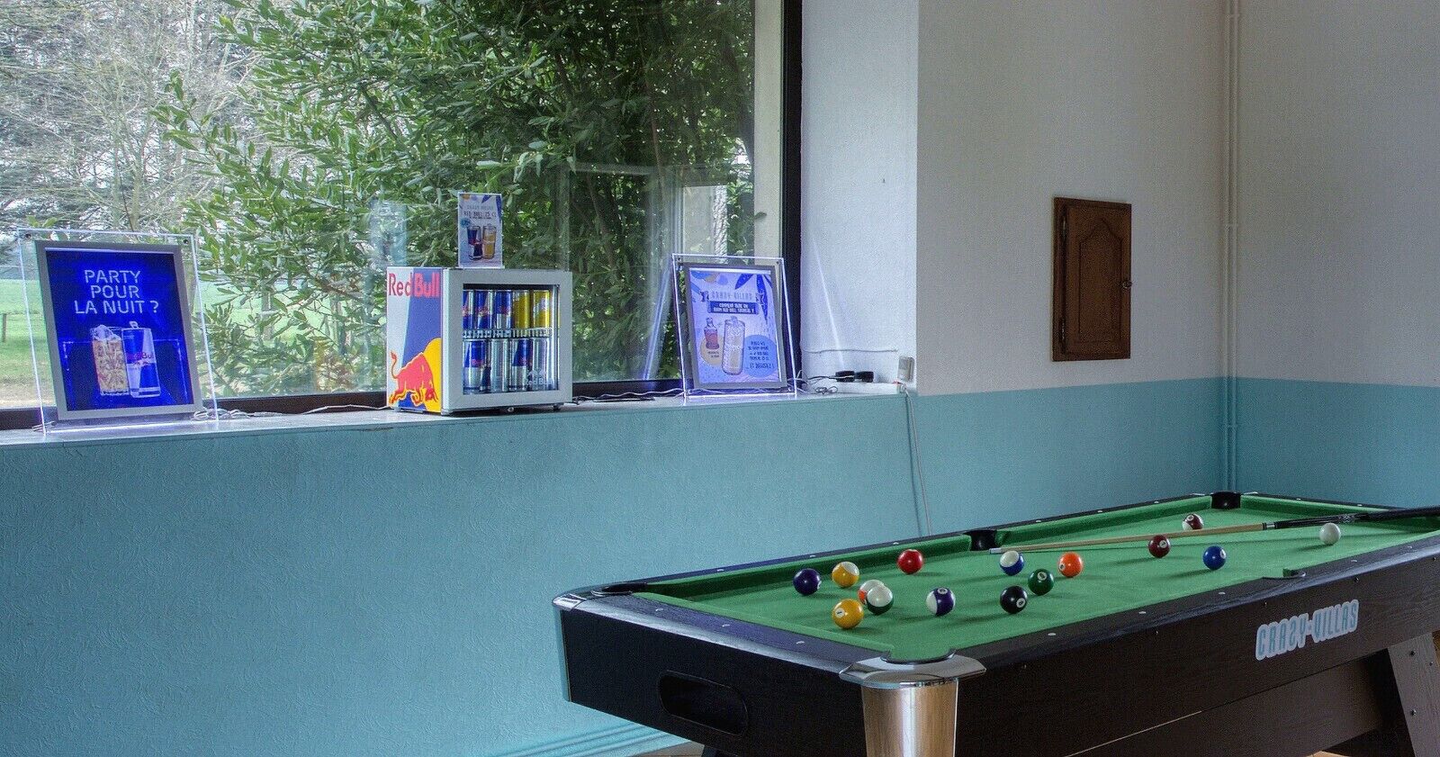 A pool table with balls, a Red Bull refrigerator, and a poster on the window sill.