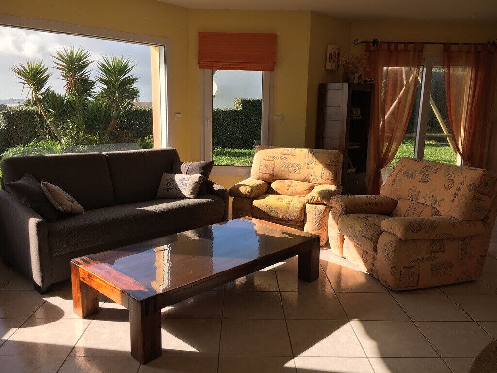 Ein Wohnzimmer mit Sofa, Sessel und Couchtisch, ein Fenster mit Vorhängen und Blick ins Grüne.