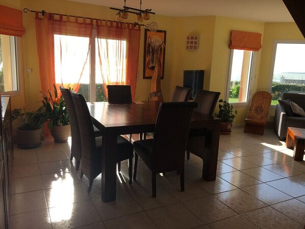 Un comedor con una mesa y sillas de madera, plantas en macetas y vistas al exterior a través de grandes ventanales.