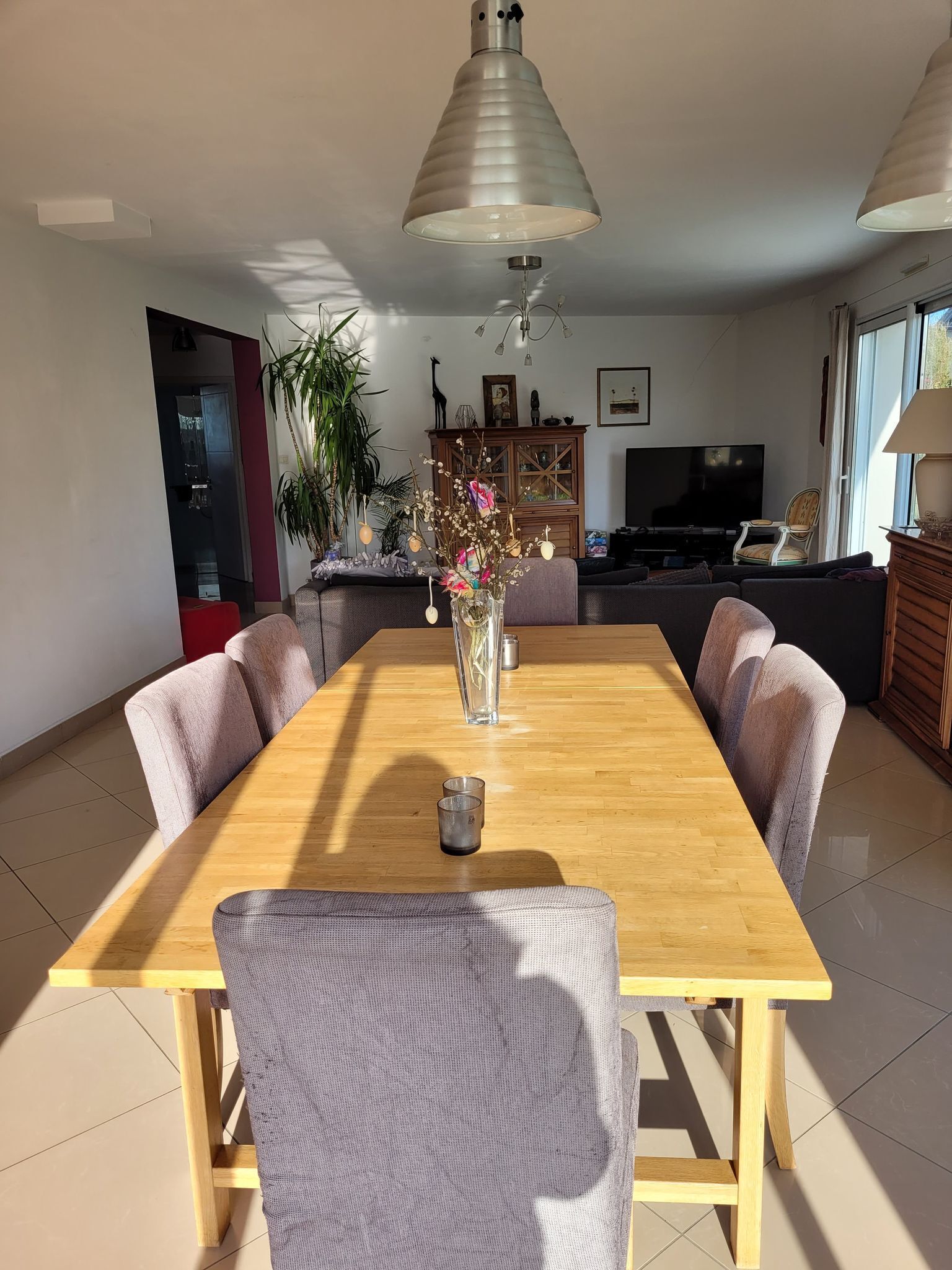 Un coin repas avec une table et des chaises en bois, un vase rempli de fleurs et un téléviseur en arrière-plan.