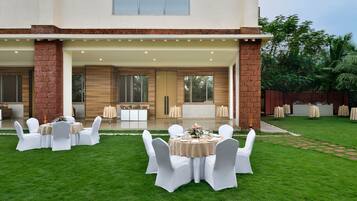 Une vaste terrasse extérieure dotée de plusieurs tables rondes et de chaises blanches, entourée d’une pelouse bien entretenue.