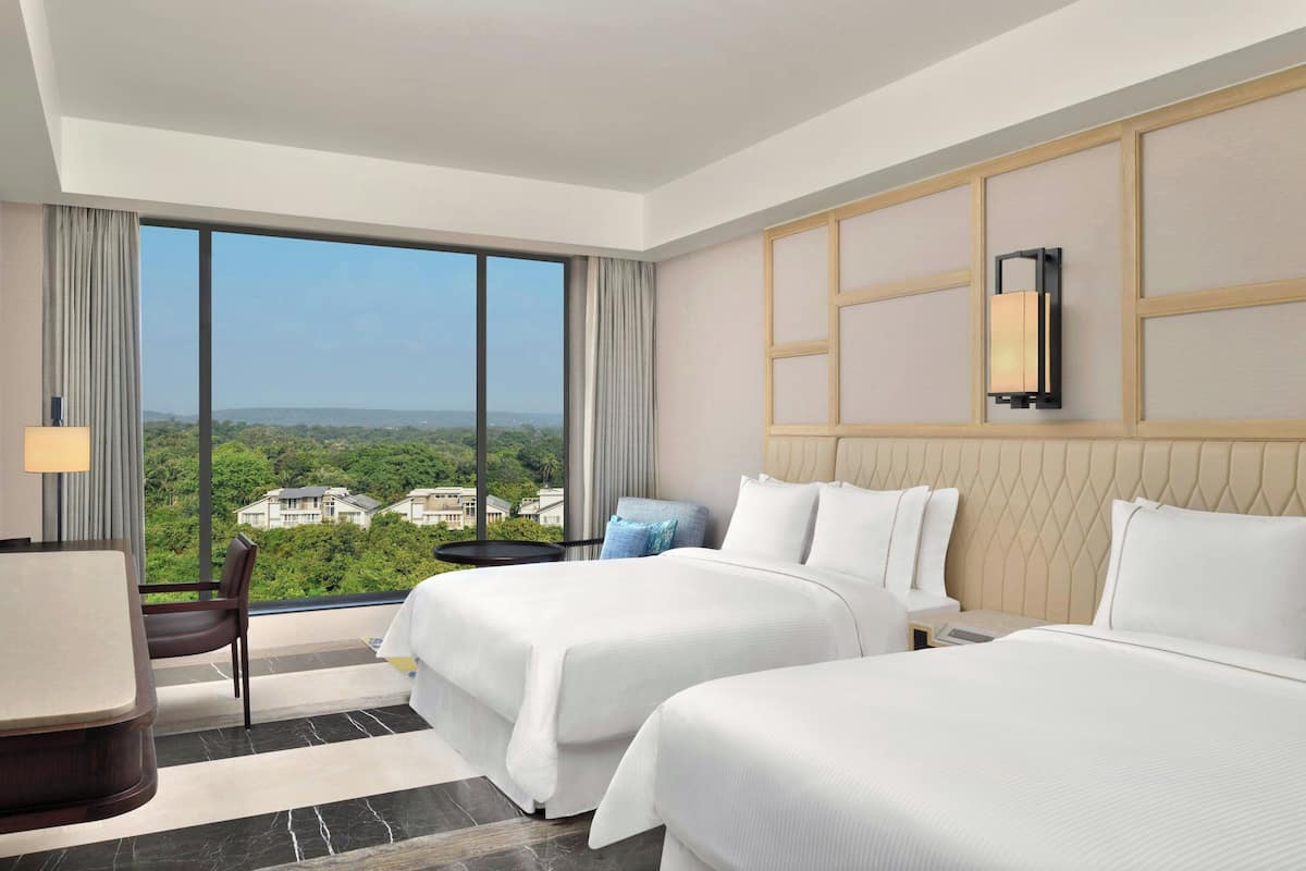 A hotel room with two beds, a desk, and a chair. There is a large window offering a view of greenery and buildings.