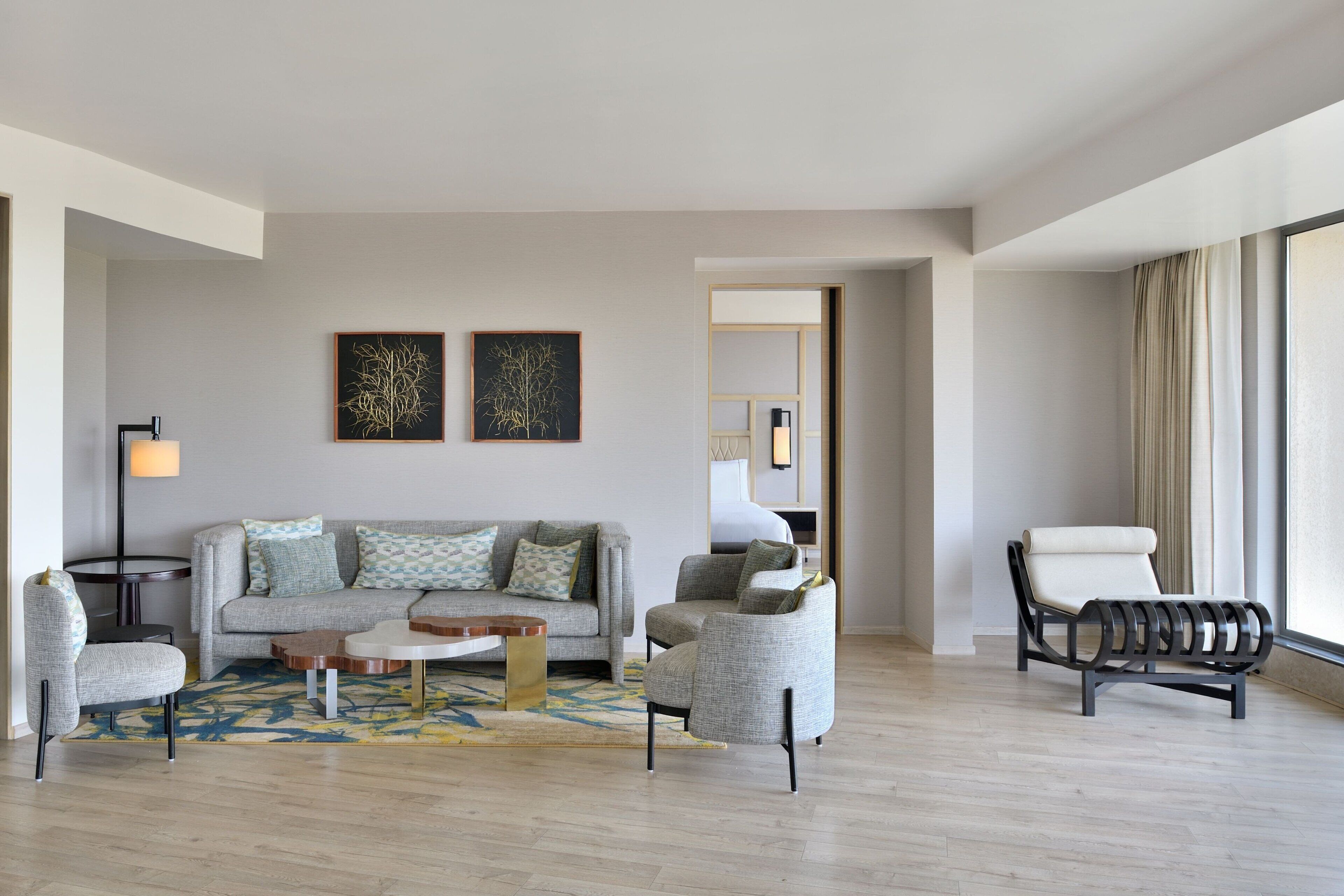 A modern living room with a grey sofa, two armchairs, a coffee table, and a floor lamp. There are two framed artworks on the wall and a large window with curtains.