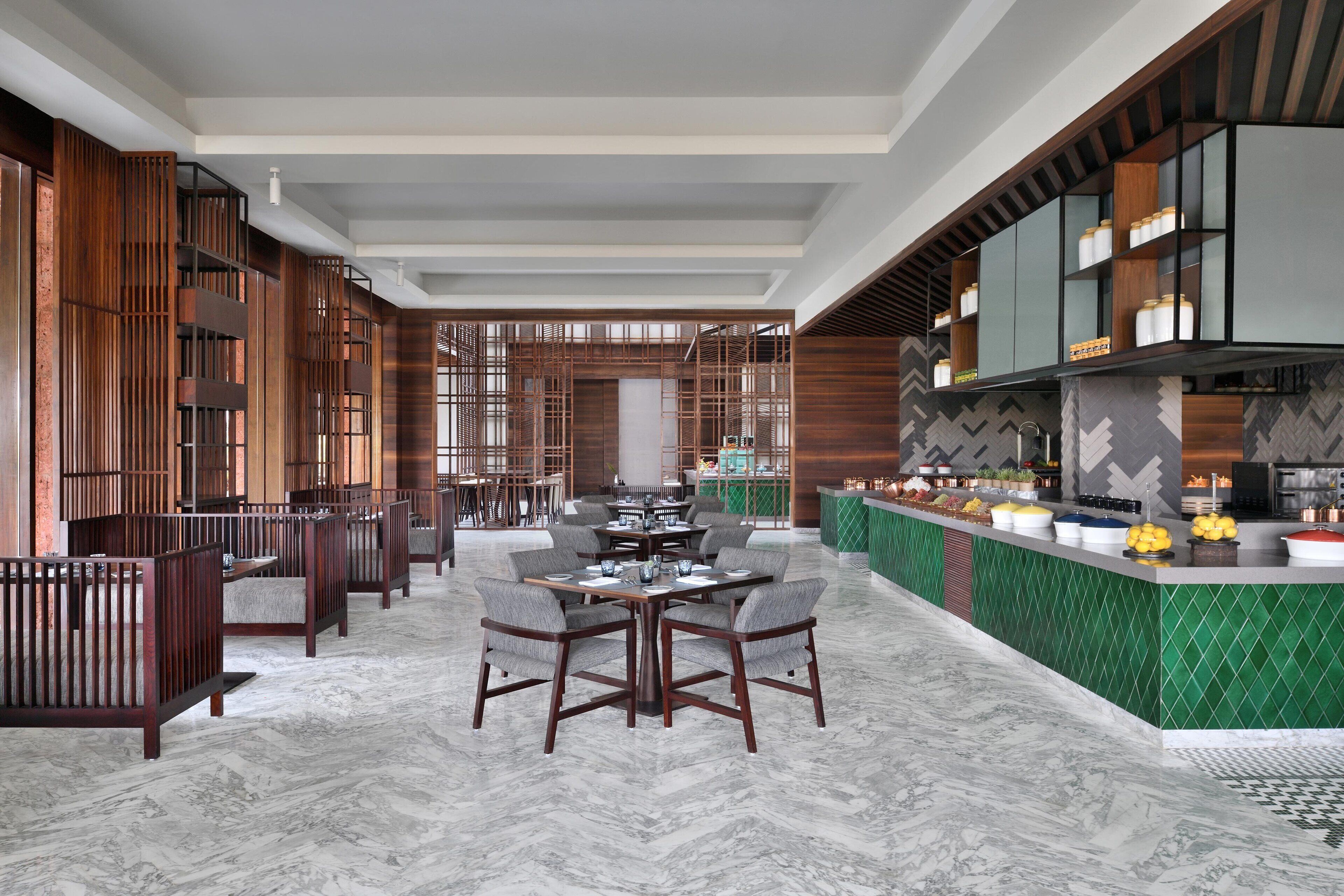 A spacious hotel lobby with a central dining area, a kitchen counter with food display, and a seating area with wooden partitions.
