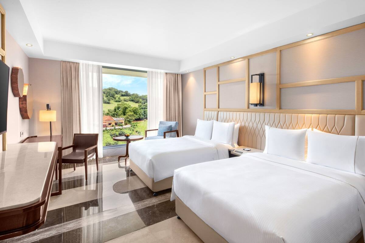 A hotel room with two beds, a desk, a chair, and a view of a green landscape.