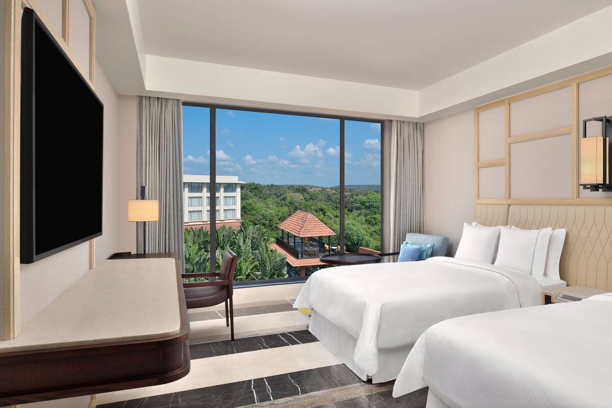 A hotel room with two beds, a desk, a TV, and a large window with a view of greenery and a building.