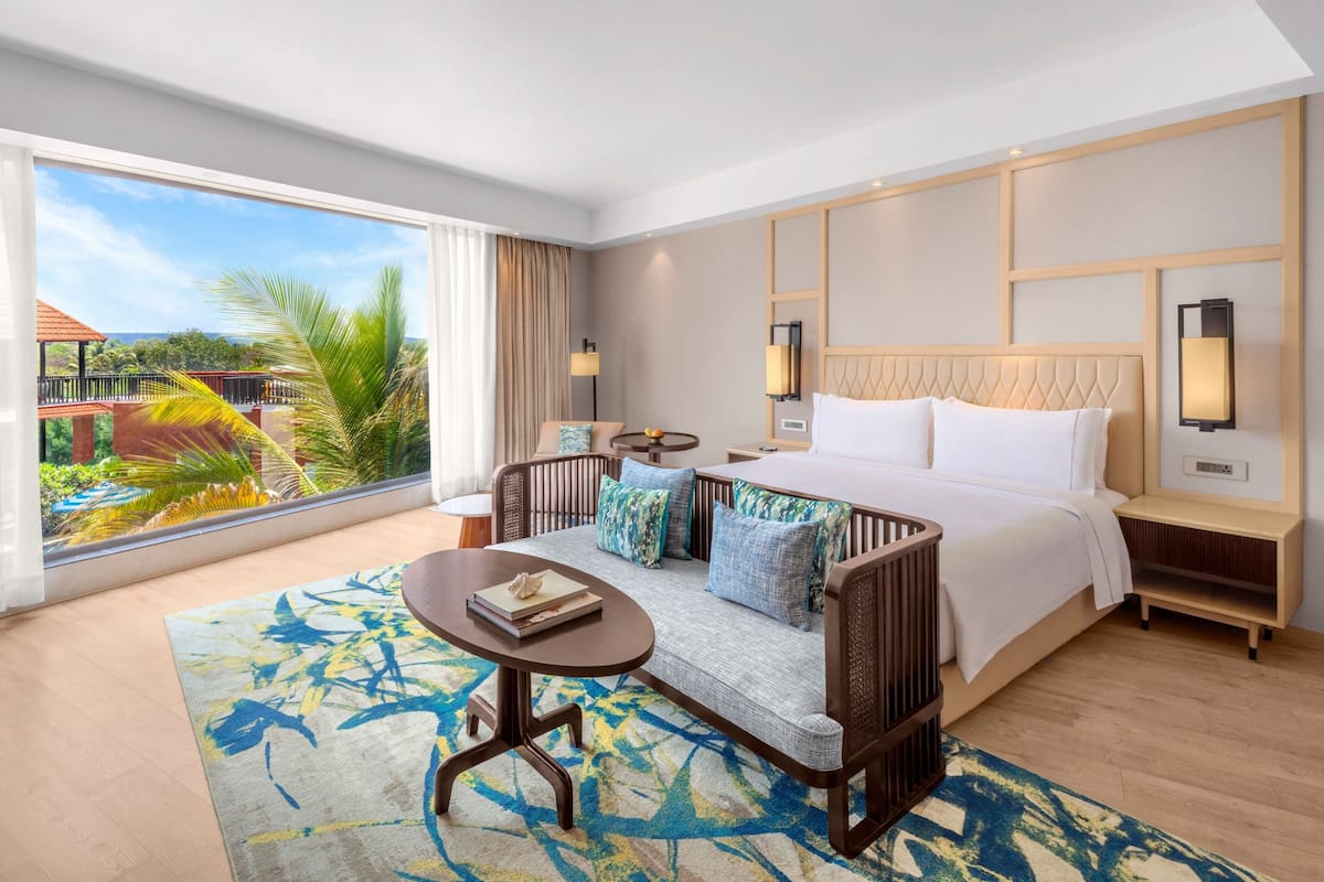 A hotel room with a large bed, a sofa, a small table, and a view of a tropical garden.