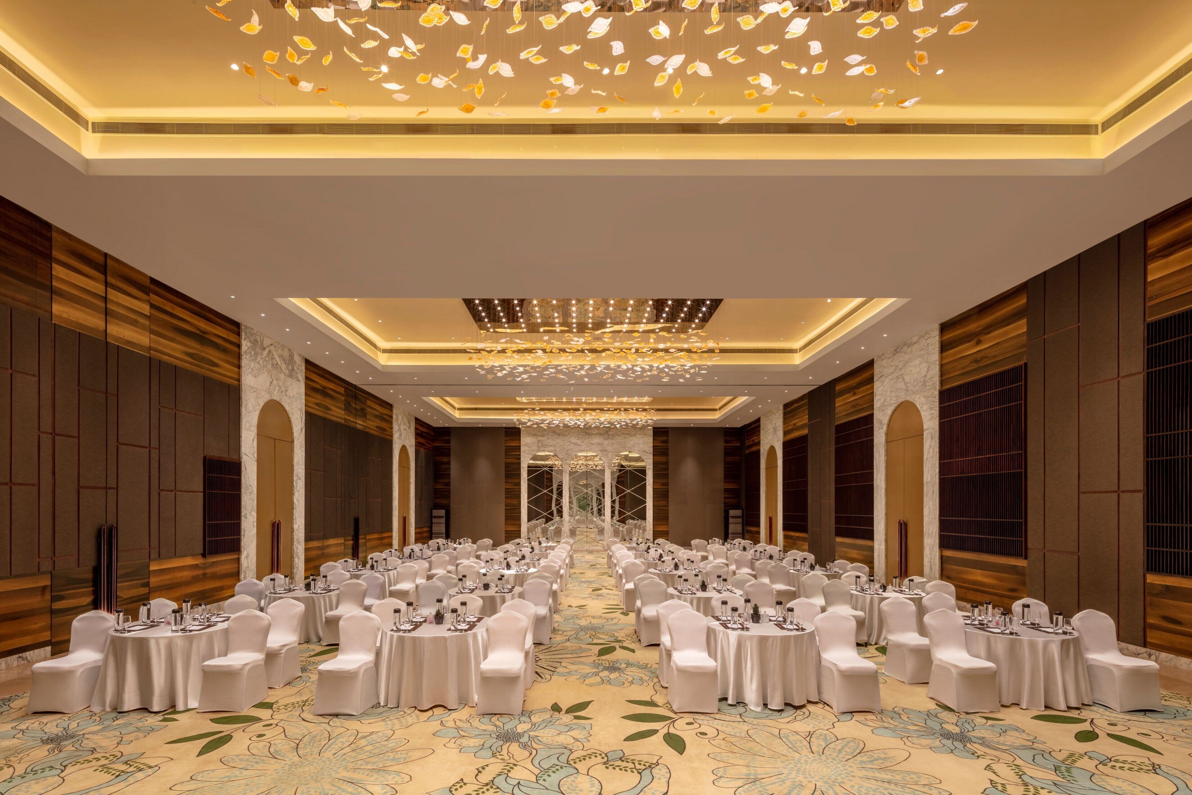 A banquet hall with round tables and white chairs, a chandelier, and a patterned carpet.