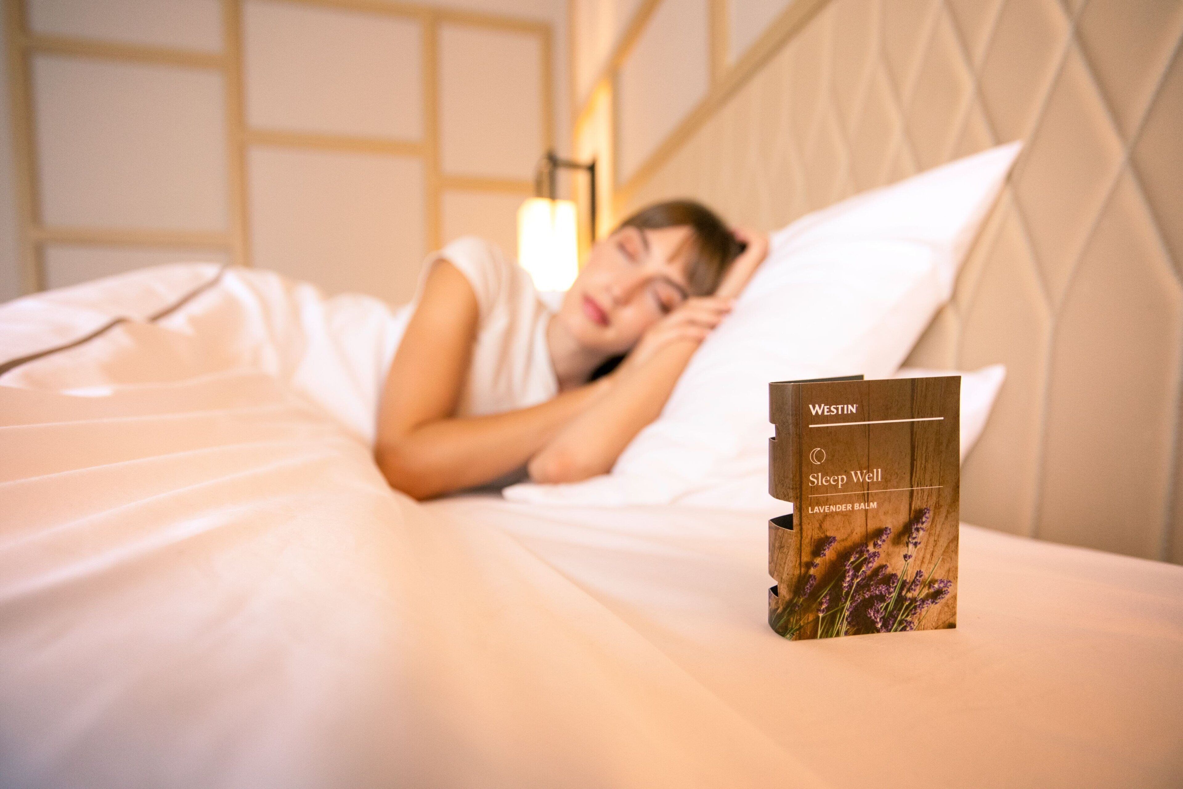 A person sleeping in a bed with a Westin Sleep Well lavender balm box on the bedside table.