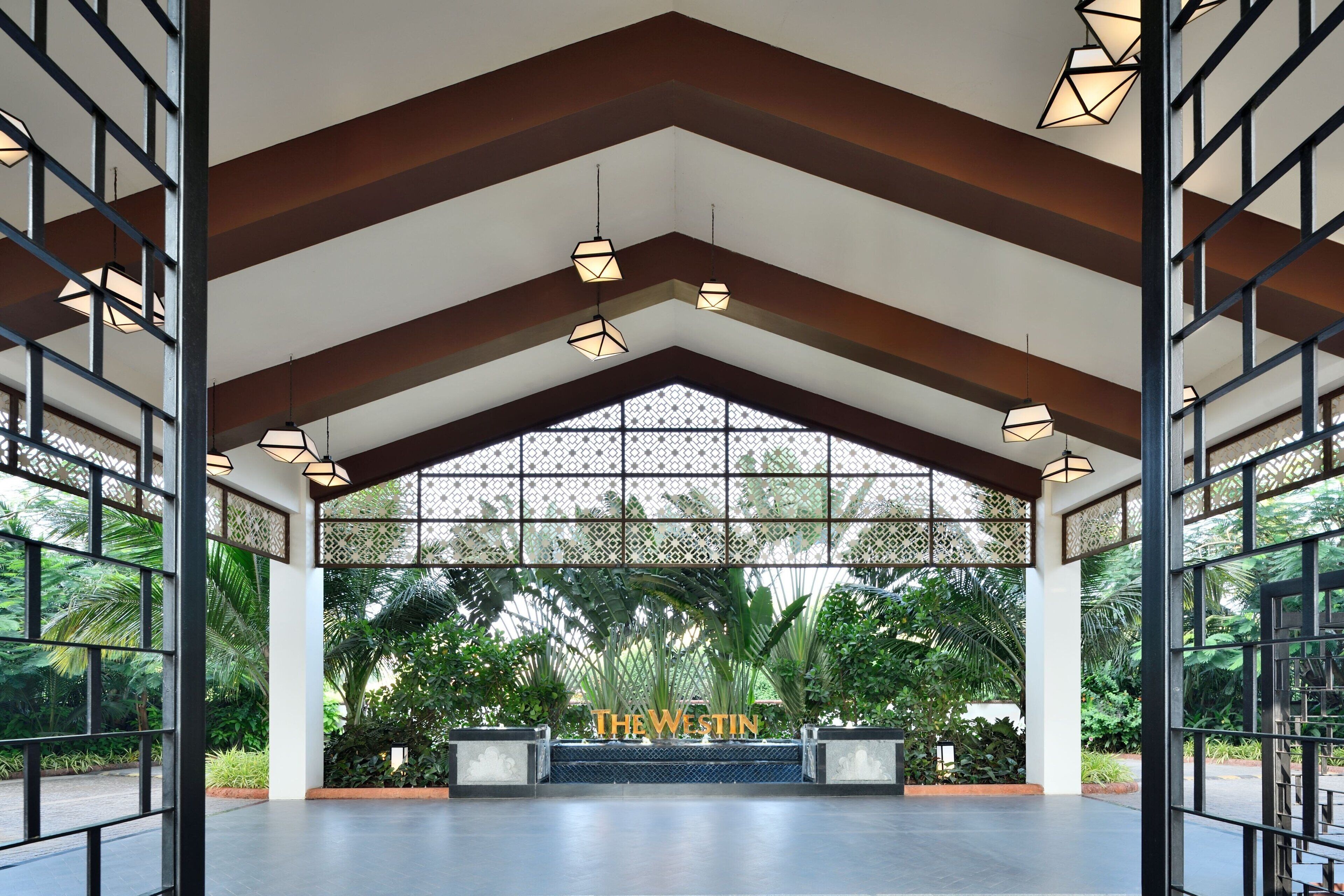A spacious lobby with a high ceiling, geometric pendant lighting, and a view of lush greenery outside.
