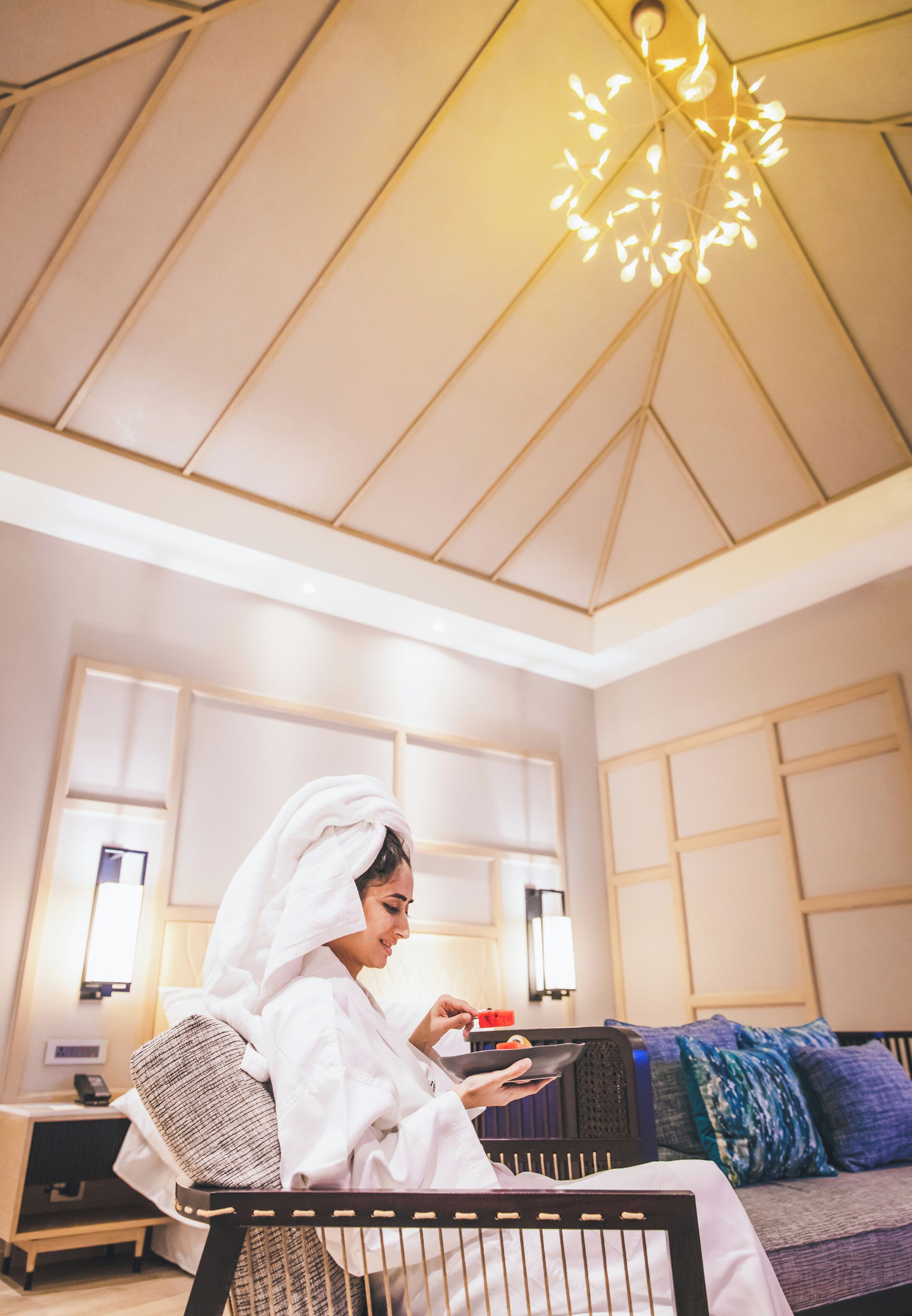 A person in a white robe sitting on a bed with a modern ceiling light, a bedside table, and a view of a room with a patterned wall and a sofa with cushions.