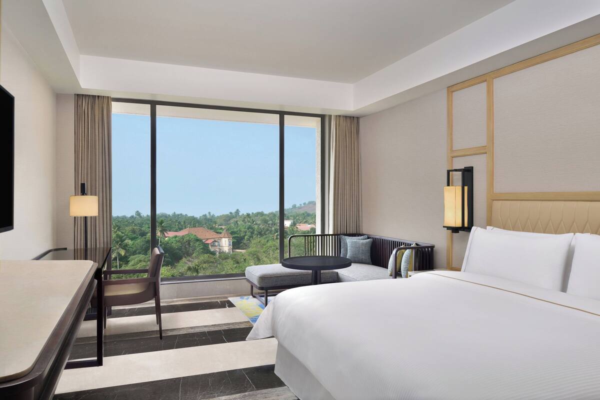 A hotel room with a large bed, a desk, a chair, and a sofa. There is a window with a view of greenery and buildings.