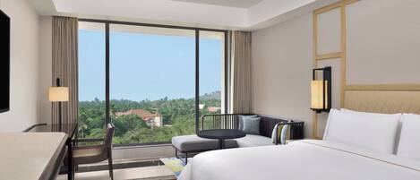 A hotel room with a large bed, a desk, a chair, and a sofa. There is a window with a view of greenery and buildings.