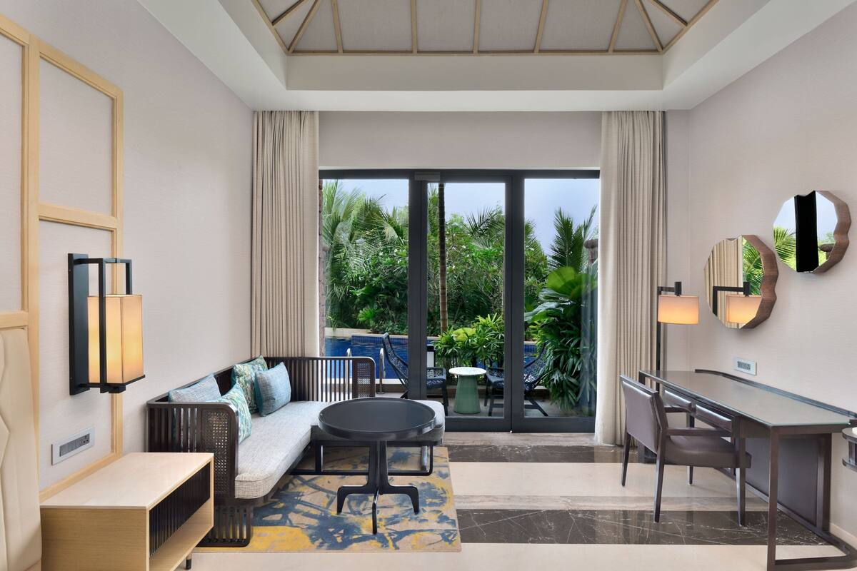 A modern hotel room with a sofa, a round table, a desk, and a view of a pool and greenery.
