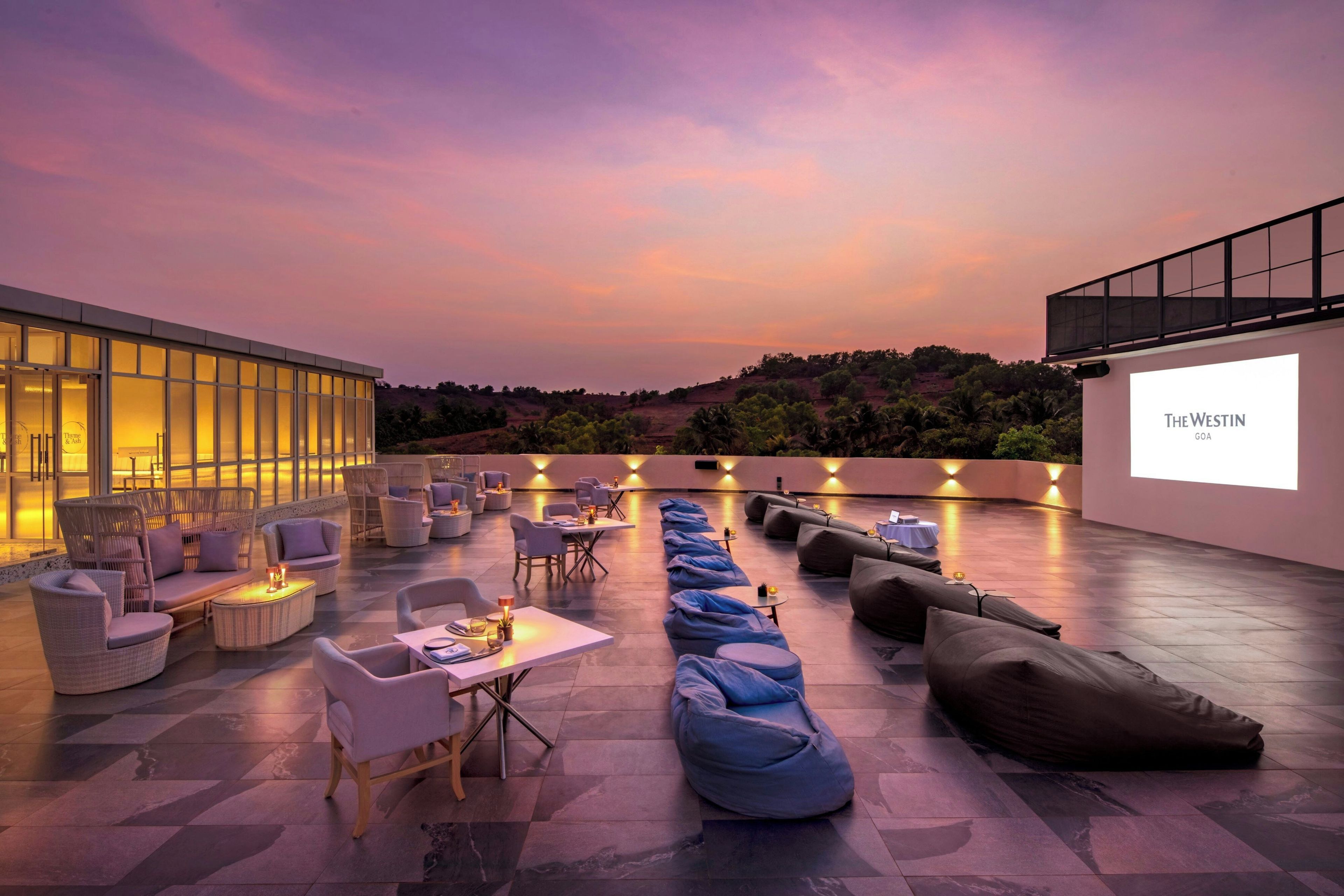 An outdoor seating area with tables and chairs, bean bags, and a projection screen displaying 'The Westin Goa'.