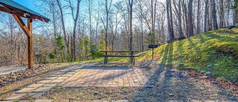 A picnic area with a bench and a shelter, surrounded by trees and a stone pathway.