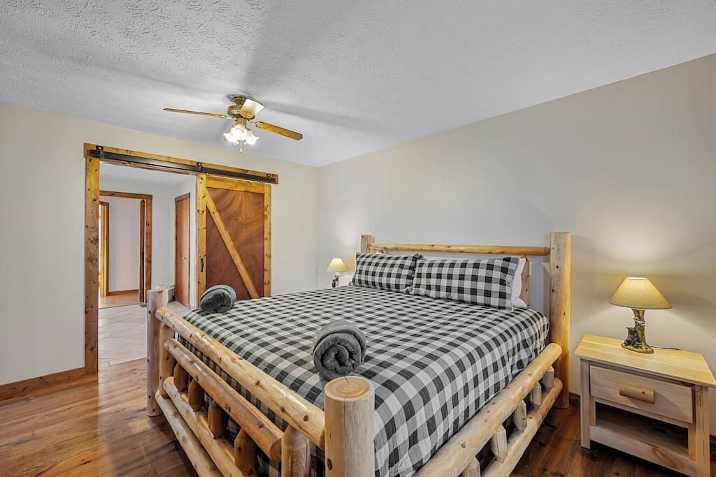 A wooden-framed bed with a checkered bedspread, a wooden nightstand with a lamp, and a ceiling fan.