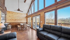 Una habitación amplia con piso de madera, un sofá de cuero, una mesa de comedor con sillas y ventanas grandes que ofrecen vistas al exterior.