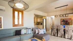Ein modernes Wohnzimmer mit einer Couch, einem Essbereich mit Stühlen, einer Küche im Hintergrund und einem großen Fenster.