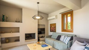 Ein modernes Wohnzimmer mit grauem Sofa, einem Holztisch und einem Kamin. Regale mit Dekorationselementen sind sichtbar, und eine Klimaanlage ist an der Wand montiert.