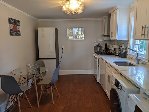 A compact kitchen with a refrigerator, stove, sink, and a small dining area with chairs and a glass table.