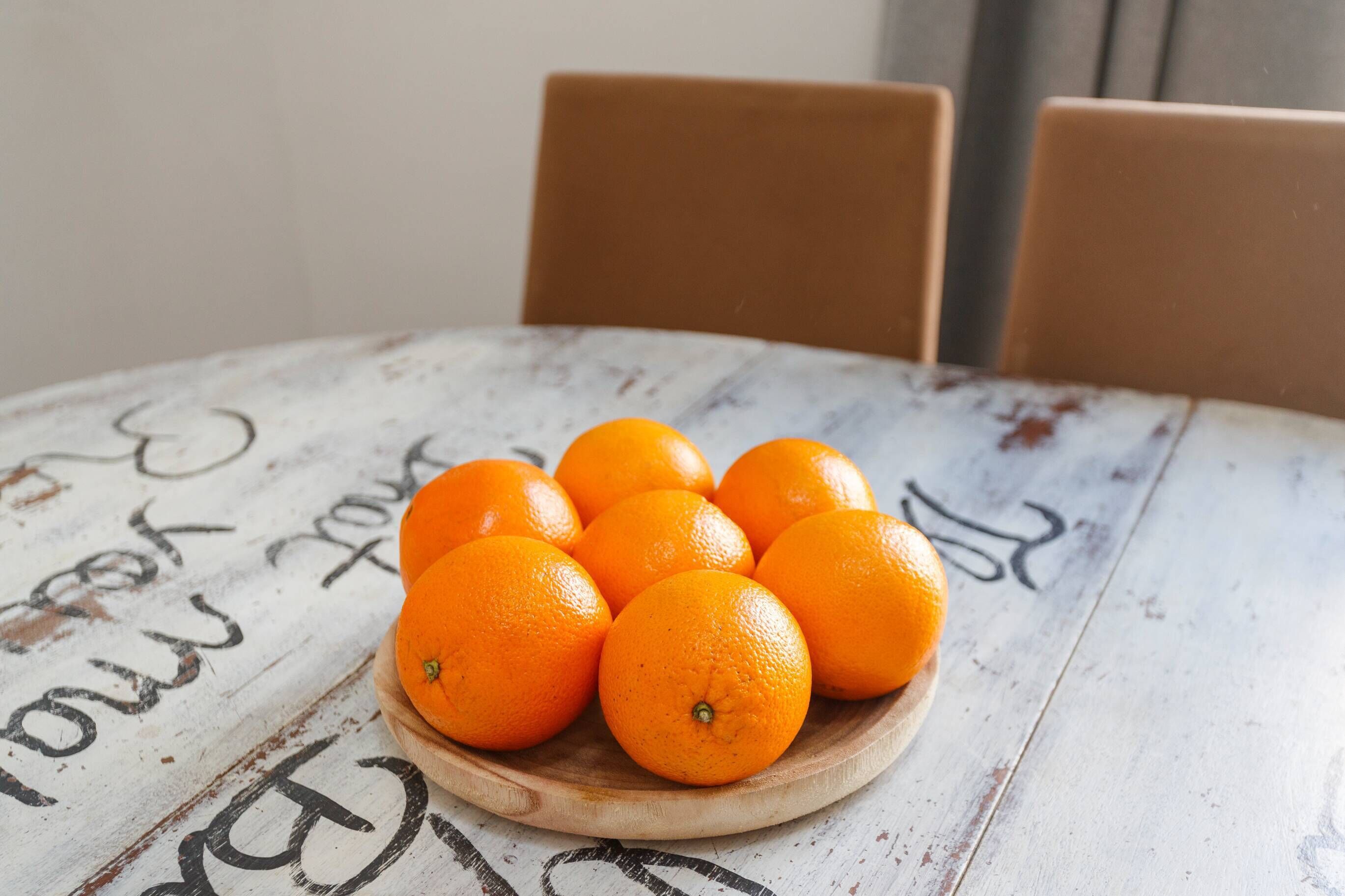 A wooden table with a white surface and black text, holding a wooden bowl of oranges.