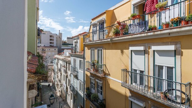 Una calle estrecha flanqueada por edificios con balcones adornados con plantas, bajo un cielo azul despejado.
