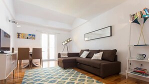 Una sala de estar moderna con un sofá seccional marrón, una mesa de comedor con sillas, un tapete estampado, una estantería y un amplio ventanal.