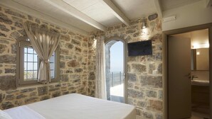 A bedroom with a stone wall, a bed, a window with curtains, a balcony, and a bathroom with a sink. - Irma Villa, a Secluded Sanctuary (Krousonas)