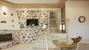 A modern living room with a stone wall, a flat-screen TV, a glass staircase, and a dining area with a glass table and chairs. - Irma Villa, a Secluded Sanctuary (Krousonas)