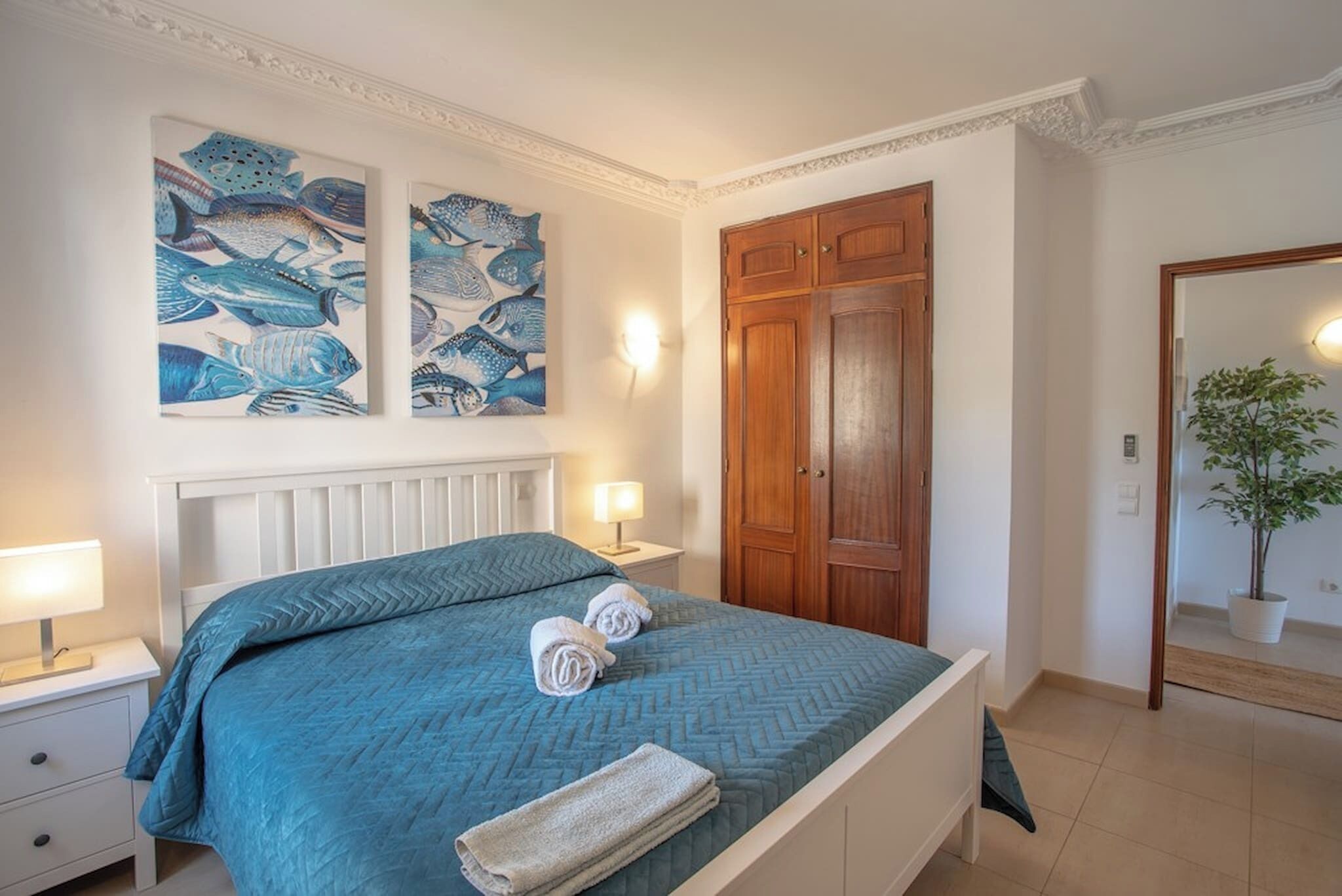Un dormitorio con cama, dos lámparas fijadas a la pared, un armario de madera y dos cuadros de peces.