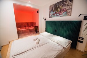 A hotel room with a bed, a green headboard, a painting on the wall, and a bathroom visible through an open door.