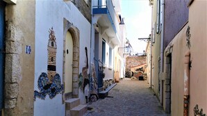 Une ruelle étroite pavée, bordée de bâtiments ornés de mosaïques décoratives et d’un mur de pierre.