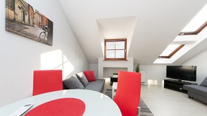 Una sala de estar moderna con techo alto, tragaluces, un sofá, un televisor y una mesa de comedor con detalles rojos.