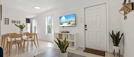 A modern living room with a dining area, a television mounted on the wall, and a door with a key lock.