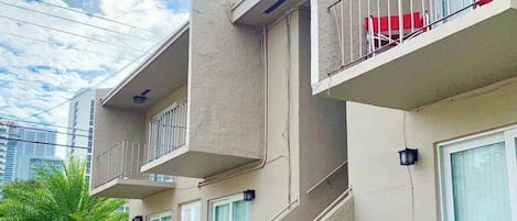 A multi-story residential building with balconies, a car parked in the driveway, and a clear sky.