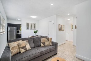 Una sala moderna con un sofá gris, una zona de cocina con un refrigerador de acero inoxidable y una mesa de comedor con sillas.