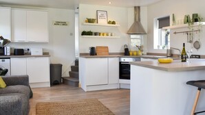 A modern kitchen with white cabinetry, a central island, and a built-in oven. There is a small seating area with a grey sofa and a brown rug. - 2 bedroom accommodation in Addingham, near Ilkley (Addingham, near Ilkley)