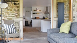 A cozy living room with a grey sofa and a wicker chair, a stone wall, and a view into a kitchen with white cabinets and a hanging light fixture. - 2 bedroom accommodation in Addingham, near Ilkley (Addingham, near Ilkley)
