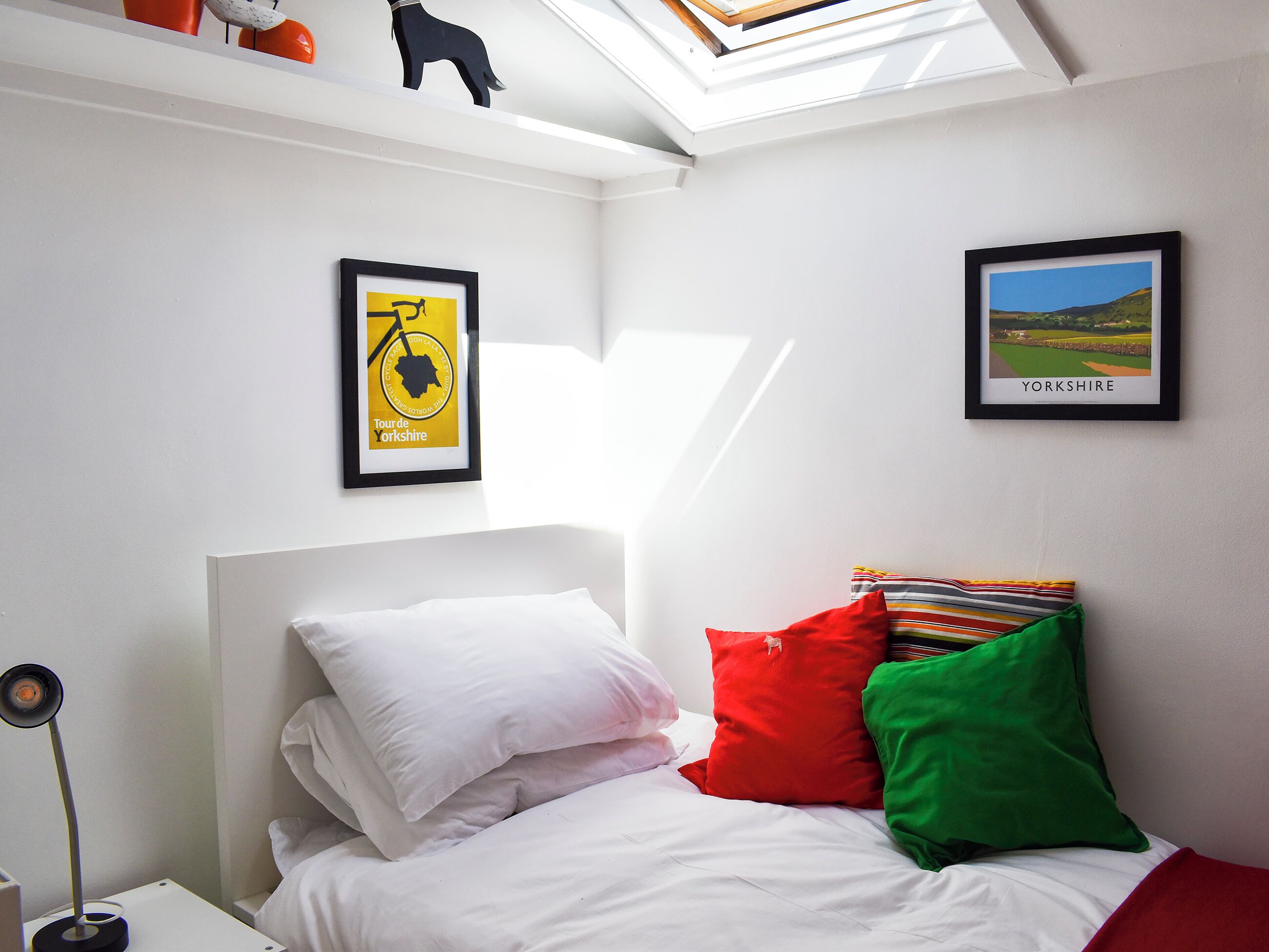 Une chambre avec une lucarne, un lit agrémenté d’oreillers blancs et colorés, et deux tableaux encadrés accrochés au mur.
