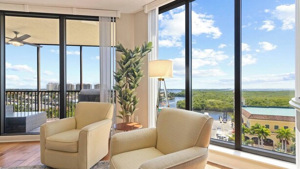 Een ruime woonkamer met twee beige fauteuils, een vloerlamp en een groot raam met uitzicht op een rivier en stadsgezicht.